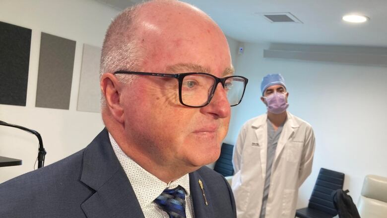 Man with silver hair and glasses wearing navy suit jacket in foreground with tall man wearing lab coat, surgical mask and cap stands in background.