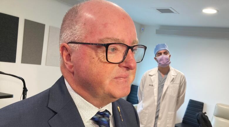 Man with silver hair and glasses wearing navy suit jacket in foreground with tall man wearing lab coat, surgical mask and cap stands in background.