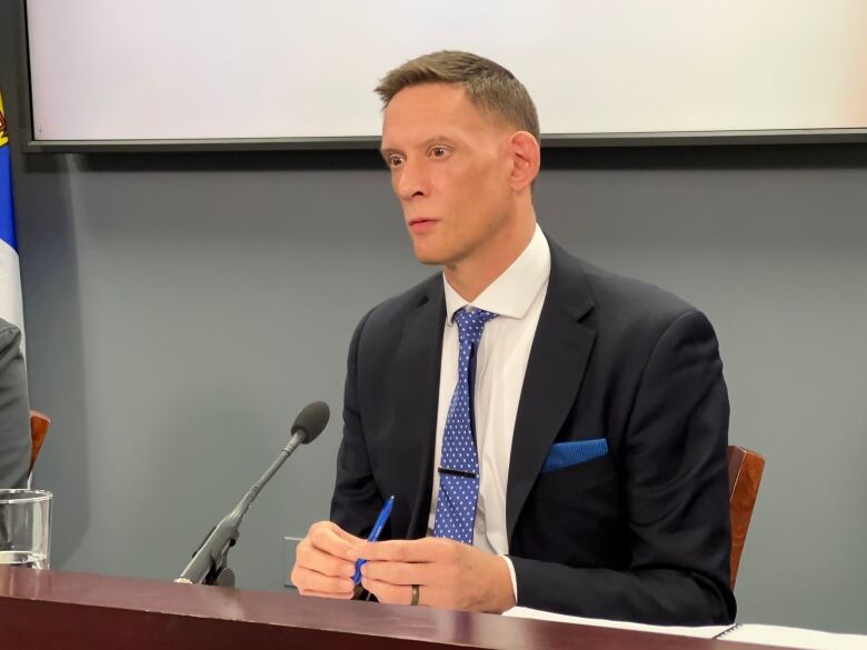 A man in a dark suit jacket, white shirt and navy blue tie speaks into a microphone.