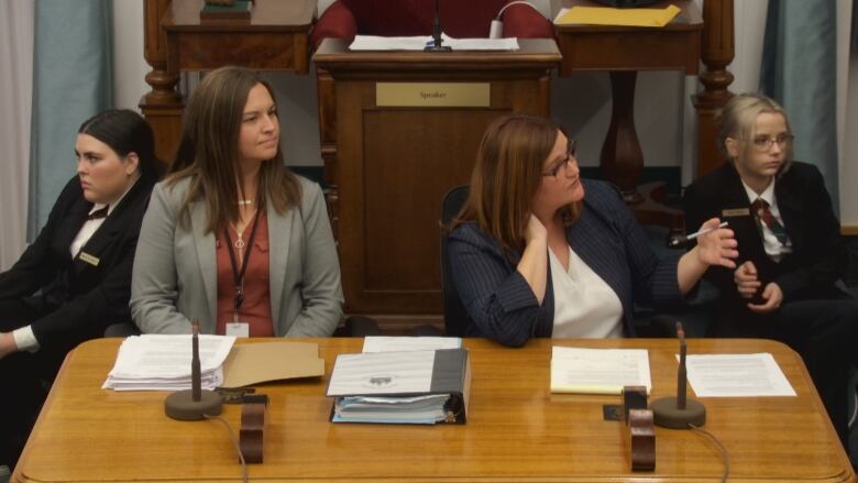 A woman in a blue jacket and white shirt sitting beside another woman, taking questions from MLAs about the sick leave bill.