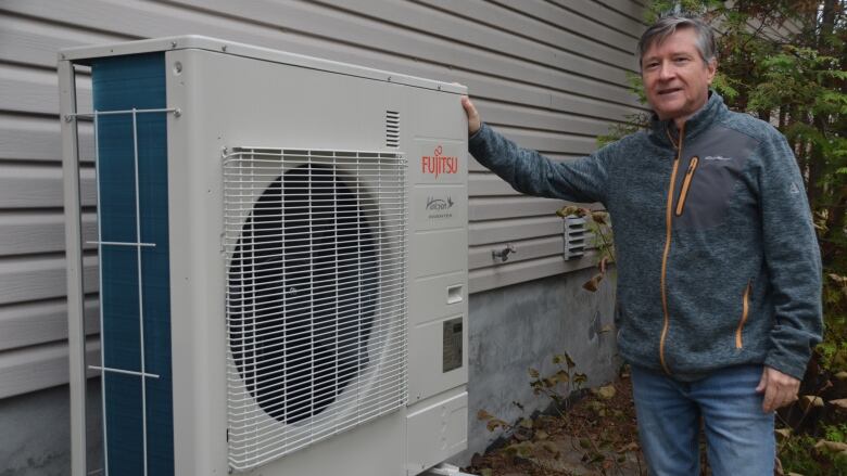 A man with grey hair, wearing a grey sweater stands next to a grey heat pump