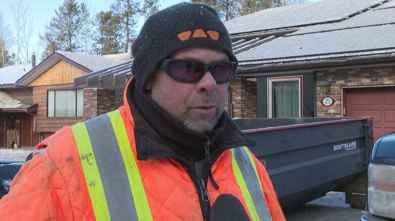 A man in a orange safety jacket, sunglasses and a toque is interviewed in front of a house.