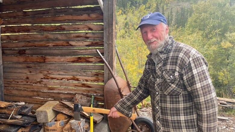 A man with a blue hat and short white beard wearing a plaid jacket. He's holding the handle of an axe that's resting on a log.