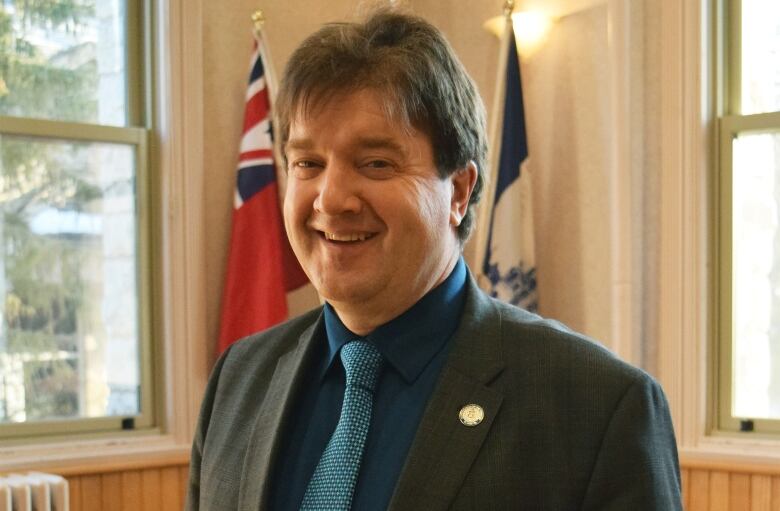 A smiling man wearing a suit stands in front of three flags. One of the flags is the Ontario flag.