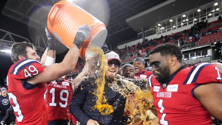 A football player empties a large gatorade cooler of orange liquid onto a laughing coach as another player looks on.