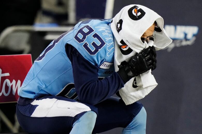 Man in a blue football uniform slumps on bench with towel on his face.