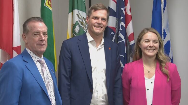 Two men and a woman - all in suits - stand next to each other smiling. They are standing indoors, in front of multiple flags.