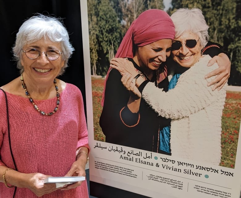 A smiling gray-haired woman stands next to a large photo of herself hugging another woman in a pink headscarf. It's a sunny day and both women are smiling brightly. 