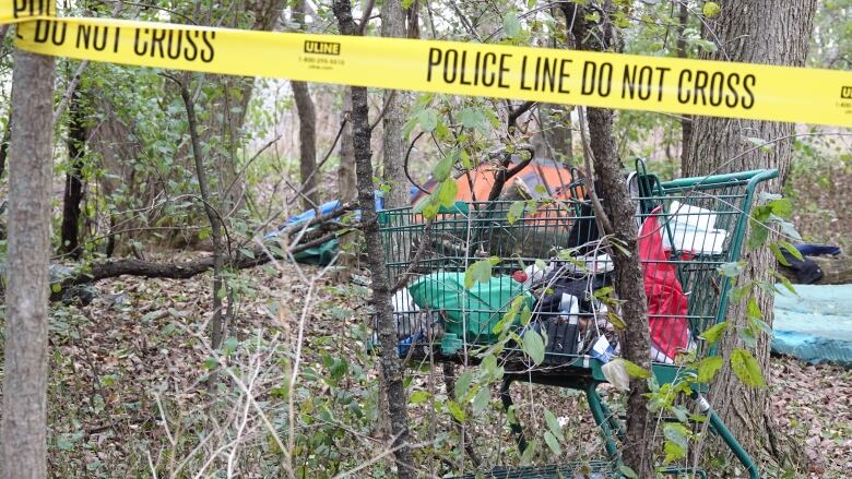 A shopping cart packed with bags and personal items is shown surrounded by trees. In the foreground is yellow police tape warning people not to cross into a crime scene.