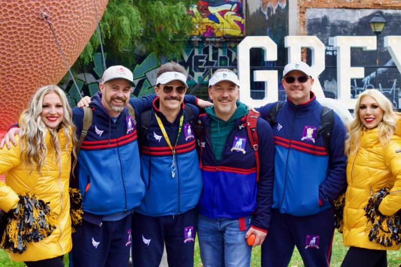 Four people in blue tracksuits and white caps post with two others who are wearing yellow winter coats and holding black and yellow pom poms. 