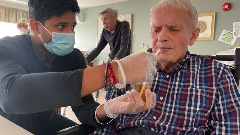 A young man wearing a face mask feeds an elderly man who is sitting at a table inside a nursing home. 