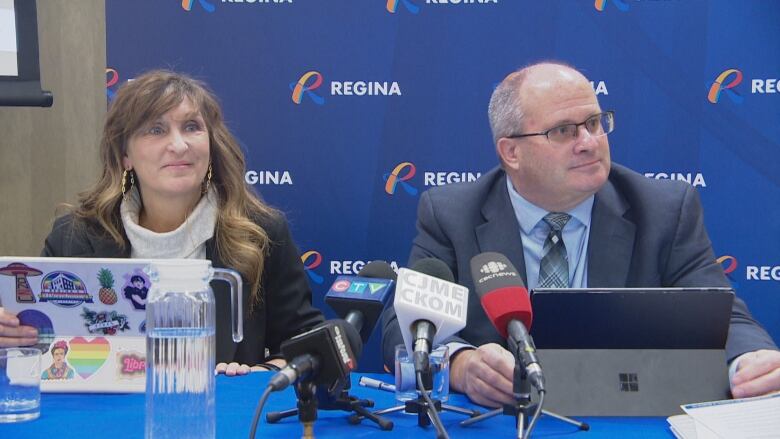 A woman with long brown hair is wearing a white turtleneck under a dark blazer. She is sitting next to a balding man with grey hair; he is wearing a blue suit and tie. The two of them are sitting at a table in front of their laptops and four microphones.