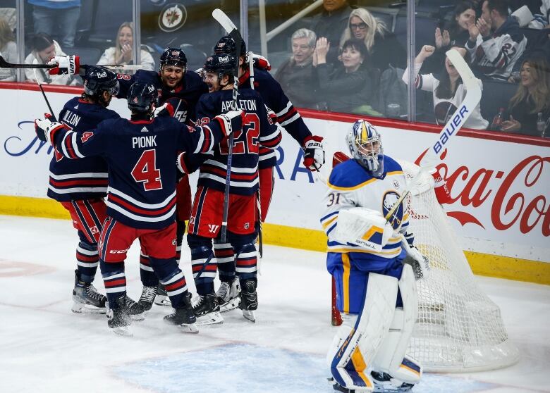 Hockey players in dark blue jerseys gather in a circle next to a goalie in a white jersey,.