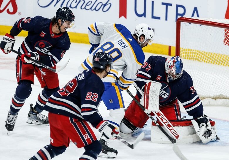 A hockey player in a dark blue jersey blocks a goal from a hockey player in a white jersey. Other players in dark blue jerseys skate around them.