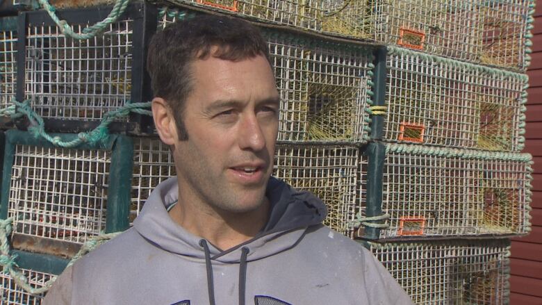 A man in a grey hoodie speaks to the camera with lobster traps behind him