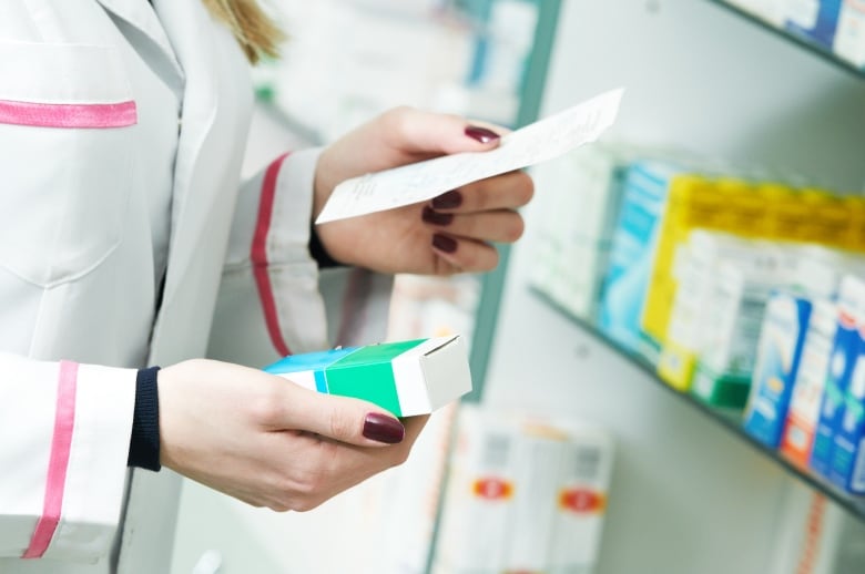 A closeup shows the hands of a woman in a white lab coat in a store, holding a prescription in one hand and a box of medicine in her other. 