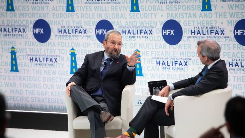 Two men wearing suits sit and speak at a conference.