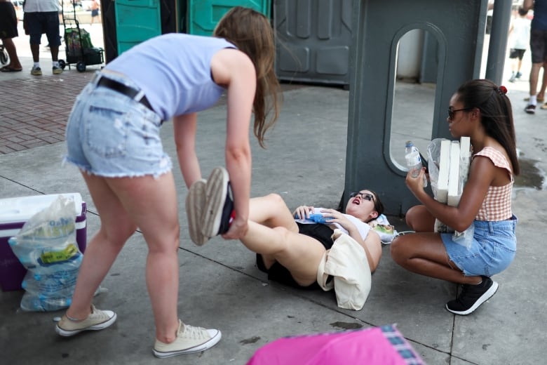 A woman bends to lift up the legs of another woman lying on the ground. A third woman kneels beside holding  bottle of water in her left hand. 