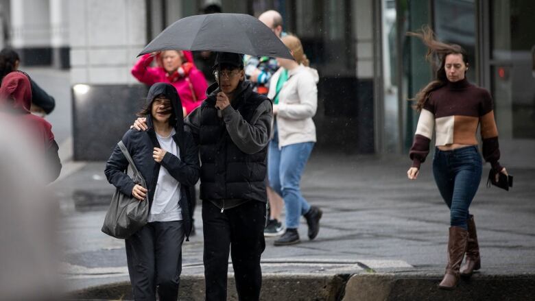 A number of women see their hair flying due to high winds in an outdoor environment, with a man trying to hold an umbrella amid the gusts.