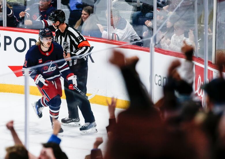 A hockey play in a dark blue jersey skates on the ice next to a referee. The audience cheers.