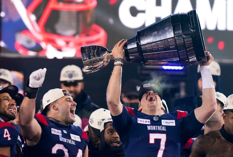 A man lifts a trophy as he screams in celebration. He's surrounded by teammates, all wearing a dark blue uniform and a white cap. 