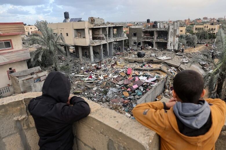 Two children stand on a balcony overlooking an urban scene where buildings are ruined and some flattened.