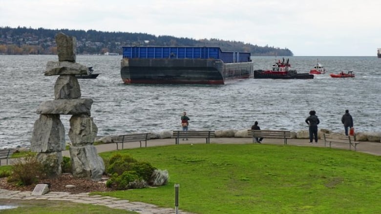 A large barge with blue siding can be seen from the shoreline where there is a large Inukshuk. People stand and watch as three smaller vessels work to push the barge away from shore.