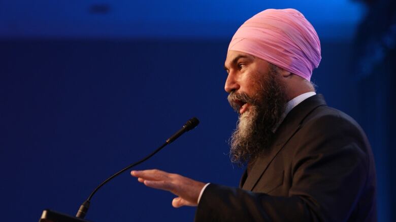 A South Asian man stands at a podium wearing a black suit and pink turban. He has a long beard and is gesturing with his hands while he speaks.