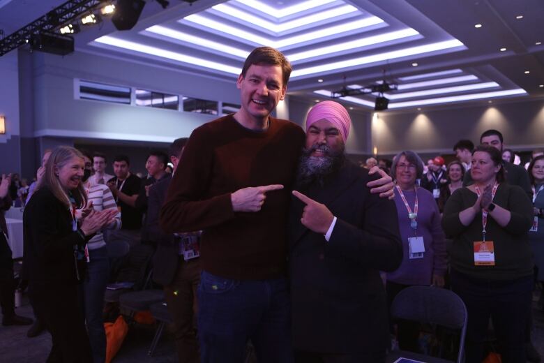 A tall middle-aged white man stands on the left wearing blue jeans and a maroon sweater. He has his left arm around the shoulders of a shorter, South Asian middle-aged man who is wearing a black suit and a pink turban. They are both smiling and are gesturing toward each other.