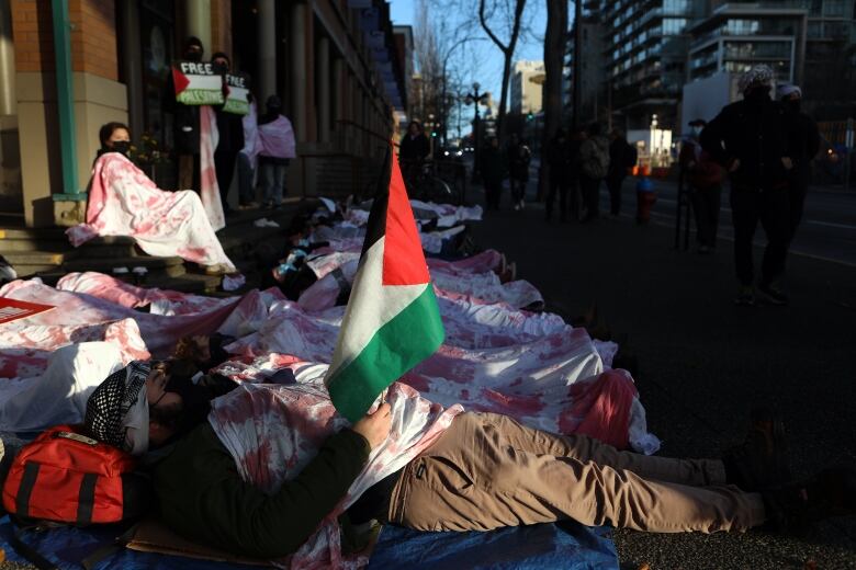 People lie on the street with sheets covering their bodies in red paint to resemble blood. One person has a Palestinian flag clutched in their hand. 