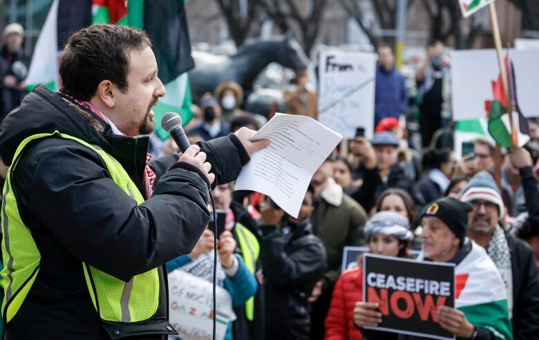 A man holds a mic and piece of paper while addressing a crowd at a pro-Palestine rally.