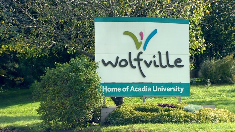 A white sign with a teal border reads 'Wolfville: Home of Acadia University' surrounded by green trees and bushes