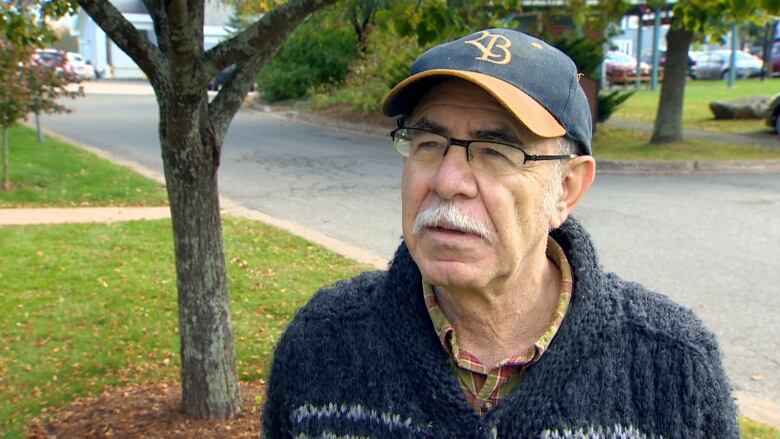 A white man with glasses and a ballhat wears a knit grey sweater while standing outside near a driveway and green park space behind him