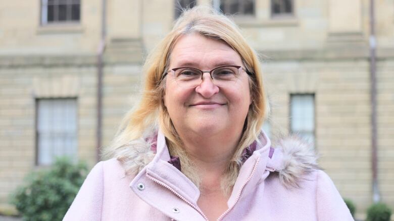 A woman with glasses stands in front of Province House in Nova Scotia. She is wearing a jacket with a hood.