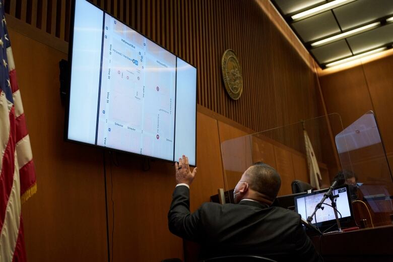 A man in a suit gestures towards a large monitor mounted on the wall. He is seated in a courtroom. 