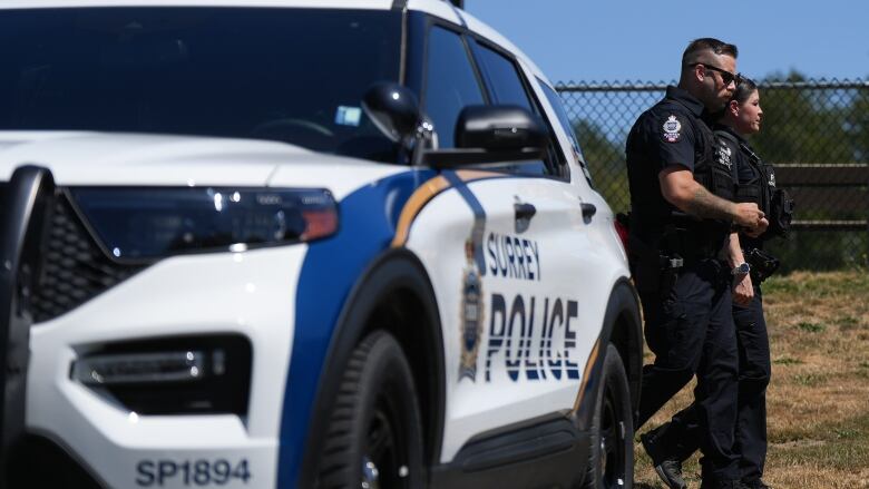 A blue and white police cruiser is pictured with Surrey Police written along the side. The car is facing forward in front of what looks like a baseball field and two uniformed officers (one male and one female) are visible walking behind the vehicle.