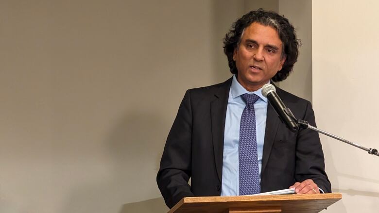 A south Asian man with long black hair, with a tuft of grey in the front, is wearing a light blue dress shirt and a blue-patterned tie under a black suit jacket. His is standing at a podium, speaking into a microphone.