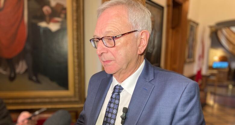 A man with grey hair and glasses addresses microphones in a hallyway.