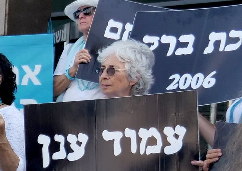 A woman with white hair and dark sunglasses hold a black placard, with white Hebrew letters, surrounded by other women holding similar signs.