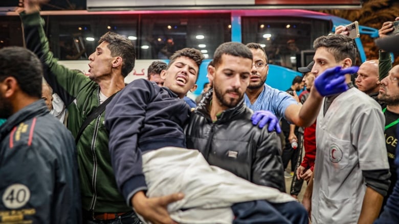 A man carries an injured teenage boy through a crowd. The boy is holding a blanket over his leg and making a pained expression. 