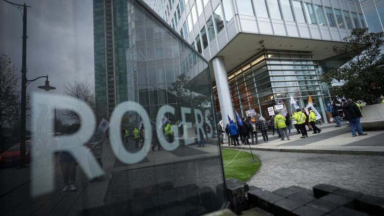 A shot of the exterior of an office building that says Rogers in etched glass. In the distance is a crowd of people holding picket signs.