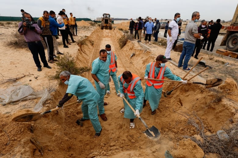 About a half-dozen men in medical-type scrubs are shown in a ditch digging as several other people and a digging machine are shown in the background.