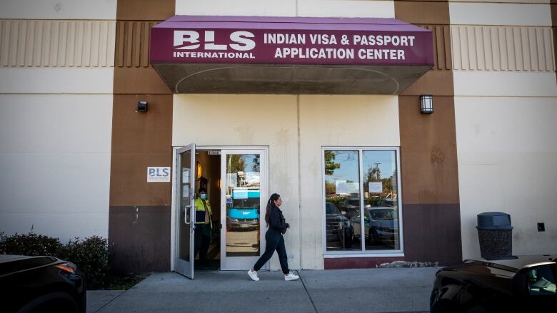 A woman walks in front of an office that has an open door. The sign above the business reads 'BLS International: Indian visa and passport application centre.' 