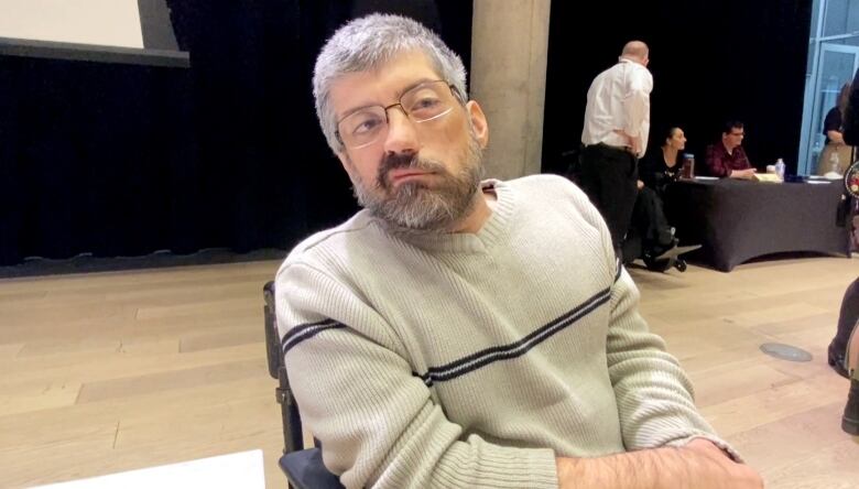 A man with glasses and a grey beard wearing a cream sweater sits in a wheelchair with a large screen behind him