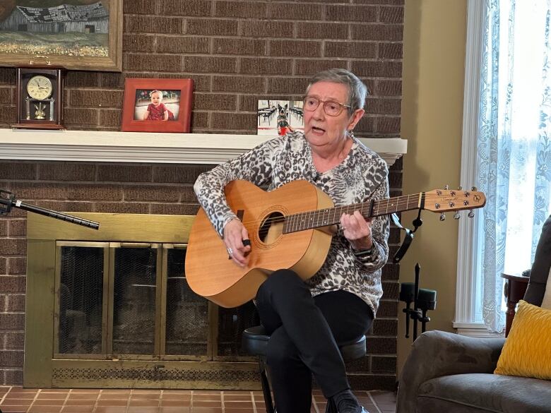 A woman sits in front of a fireplace playing the guitar.