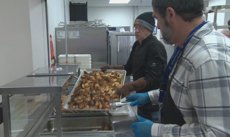 Two men work at a kitchen, one of them holding a tray of cooked potatoes.