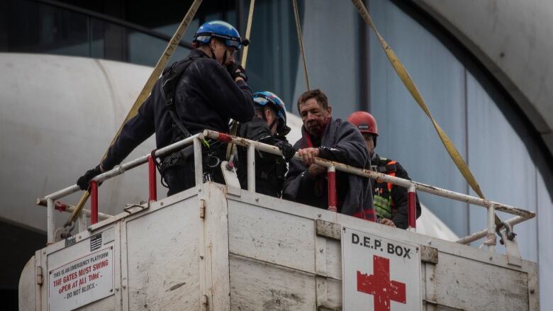 An injured man with blood on his face is seen with three rescuers in a box being lowerer by a construction crane. 