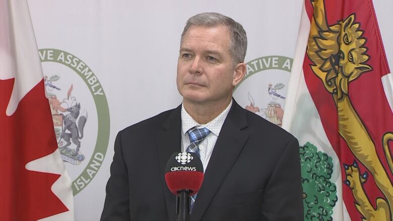 A man in a dark suit and white shirt standing in front of a P.E.I. Legislative Assembly background taking questions from reporters.