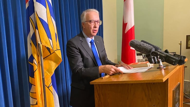 A white male with white hair wearing a black suit and blue tie stands at a podium.