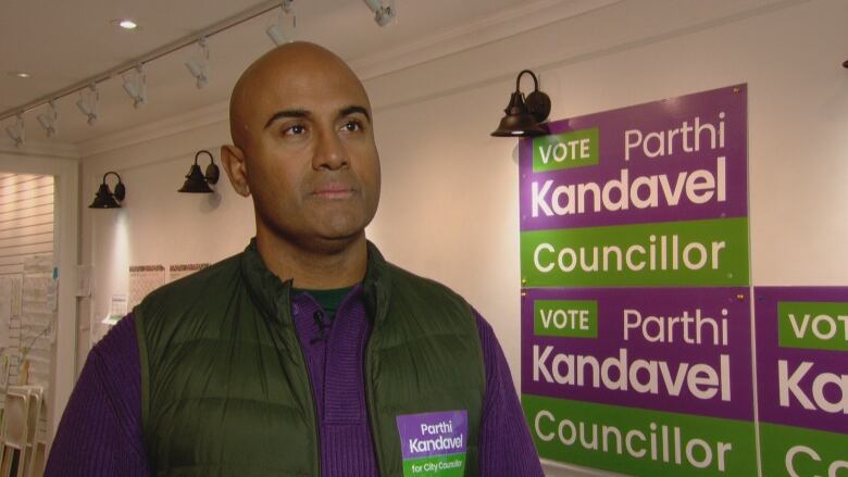 Man in a green vest and purple shirt stands in front of campaign signs.